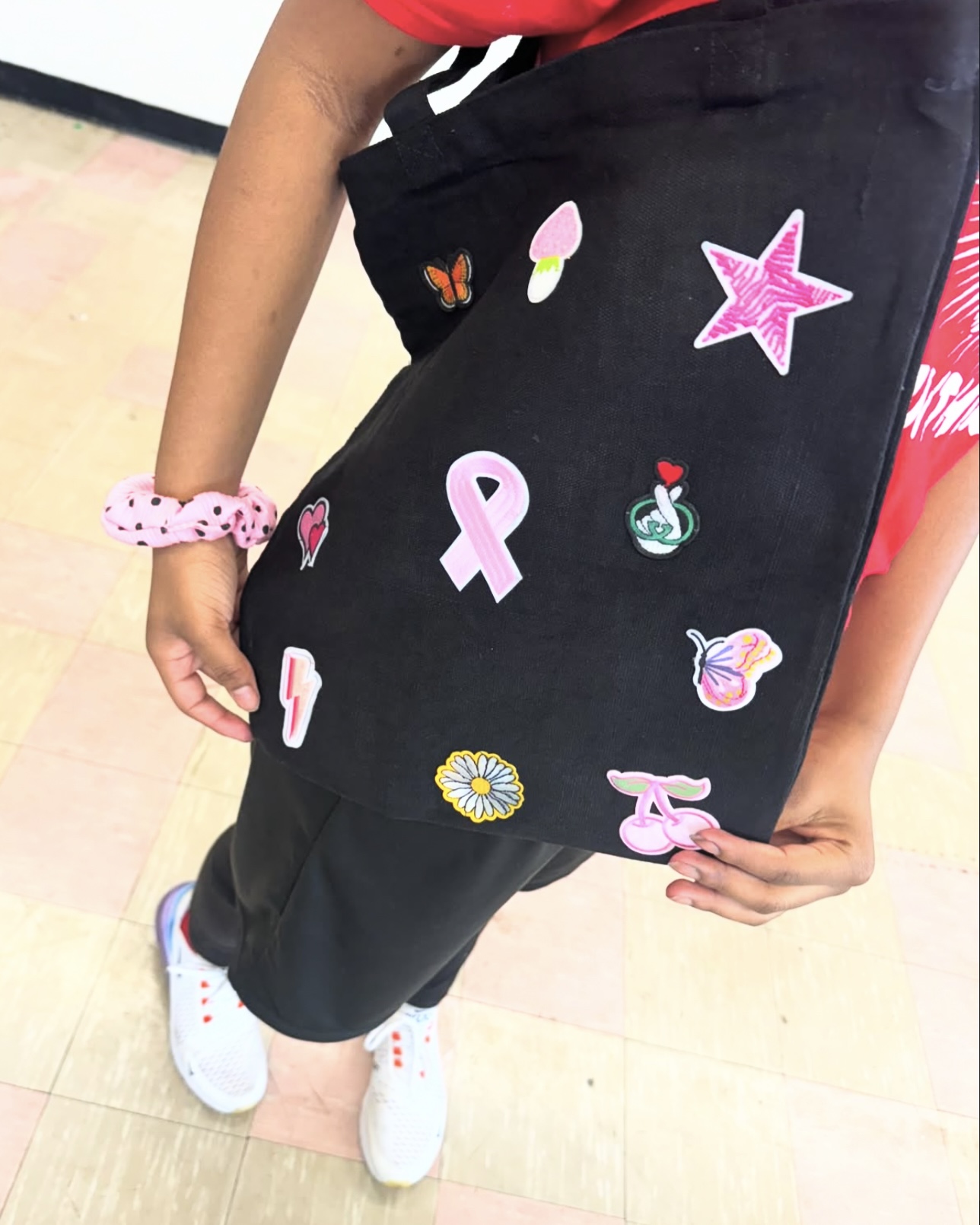 Person holding a black bag decorated with colorful patches, wearing a pink scrunchie, white sneakers, and a red shirt.