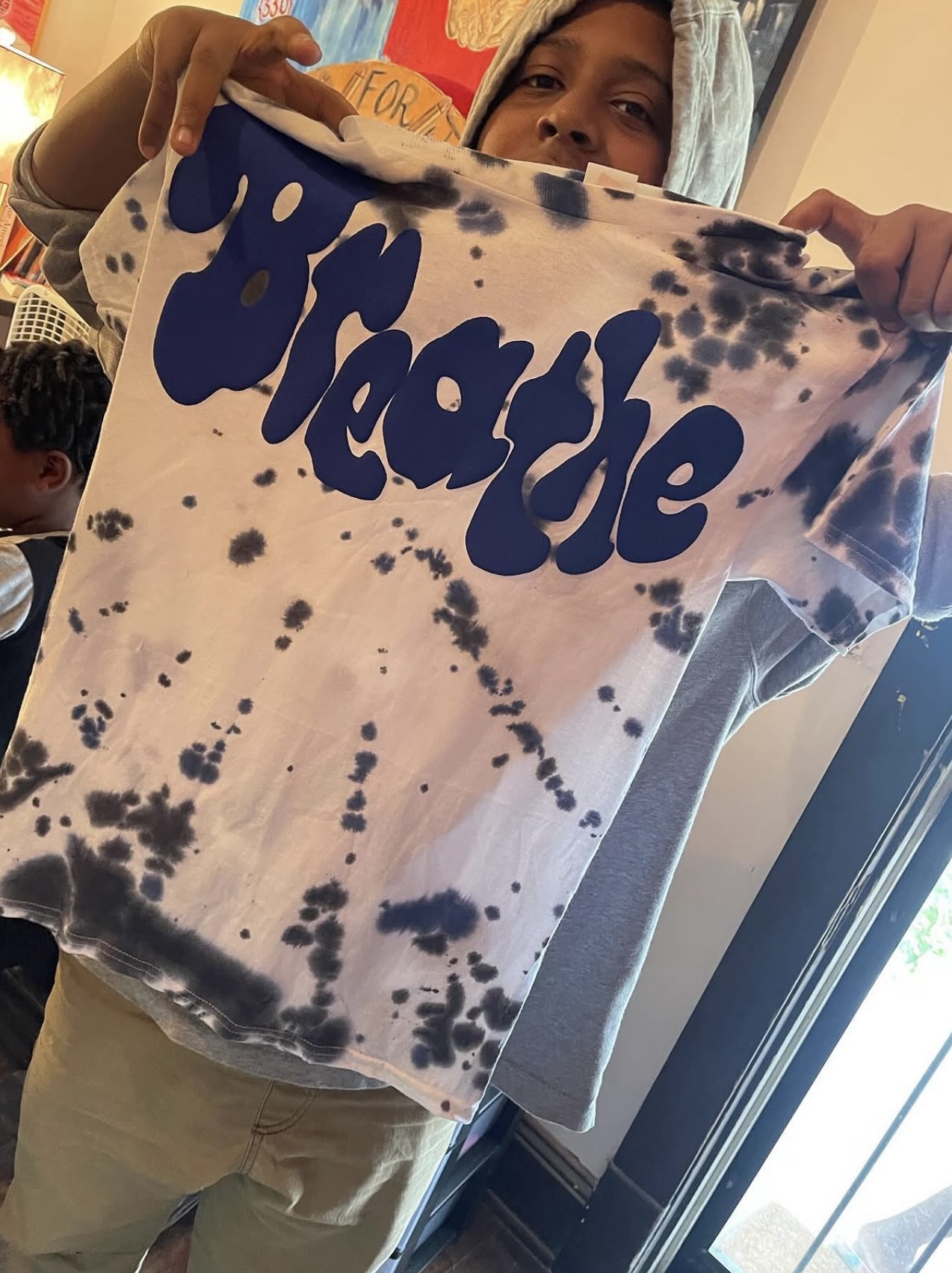 Person holding a tie-dye bag with the word 'Wicked' in blue, partially covering their face, near a window.