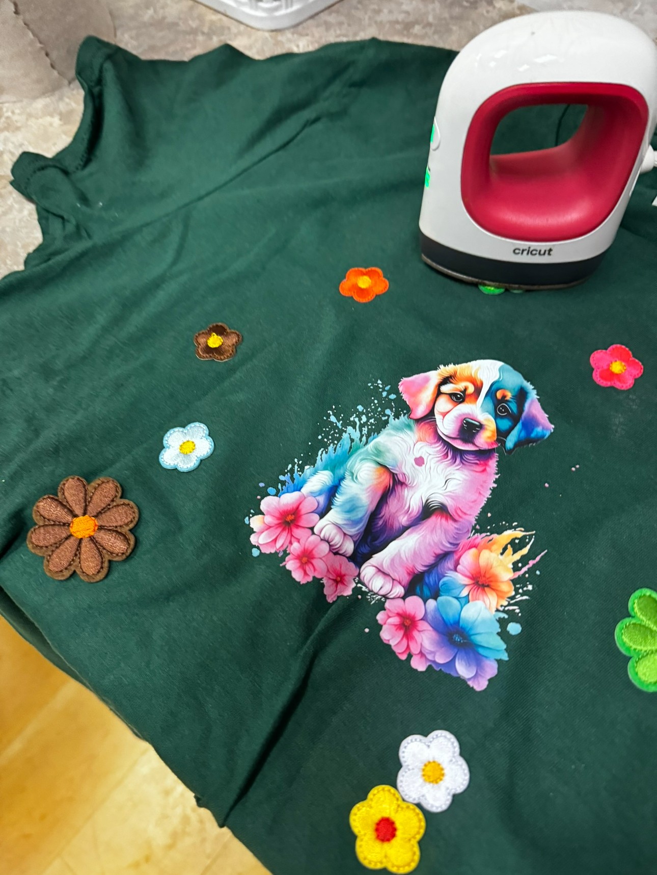 Green fabric with colorful flower decorations and a puppy illustration, iron, and other objects on top.