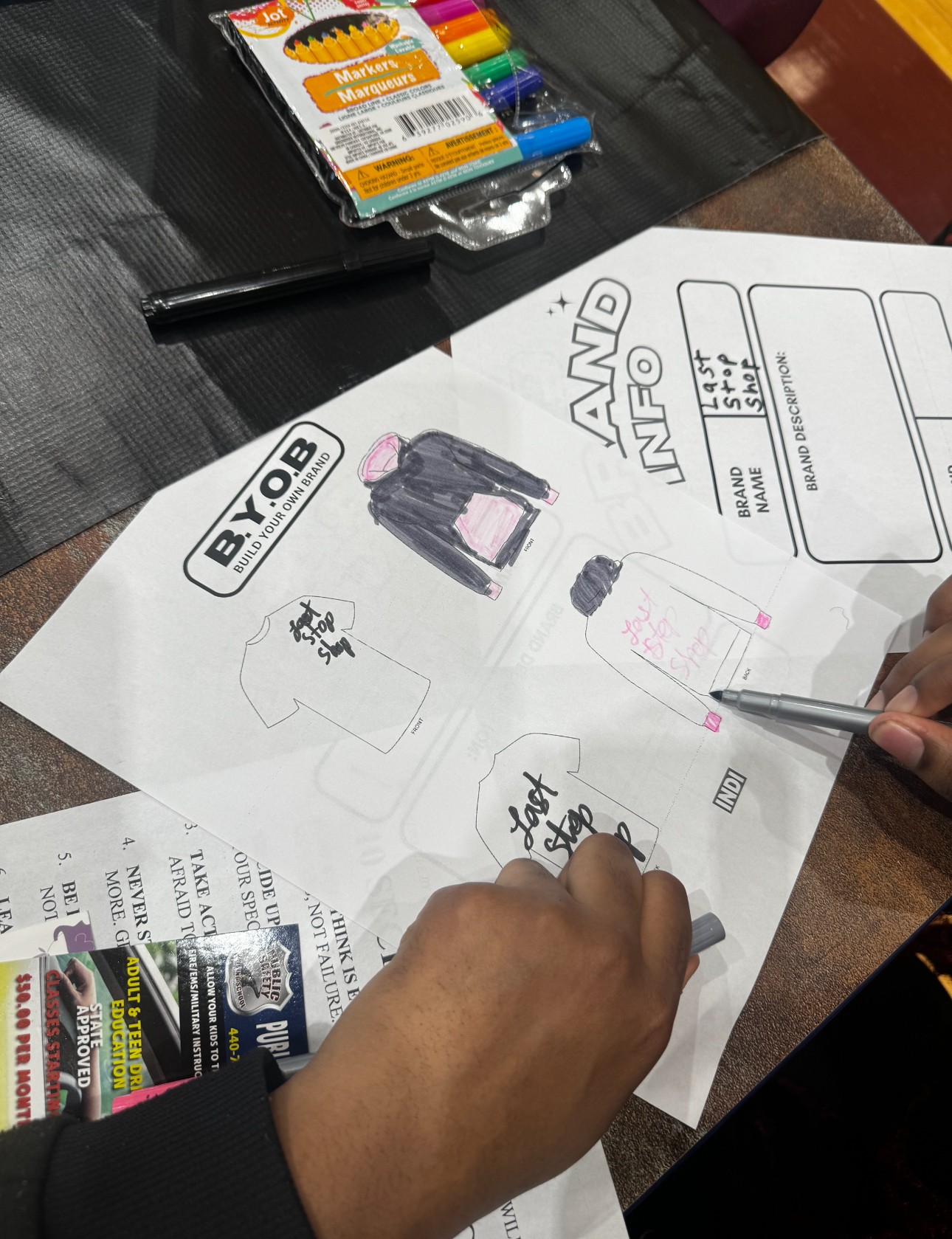 Person drawing on a large sheet of paper with various sketches and text, on a dark table with colored markers nearby.