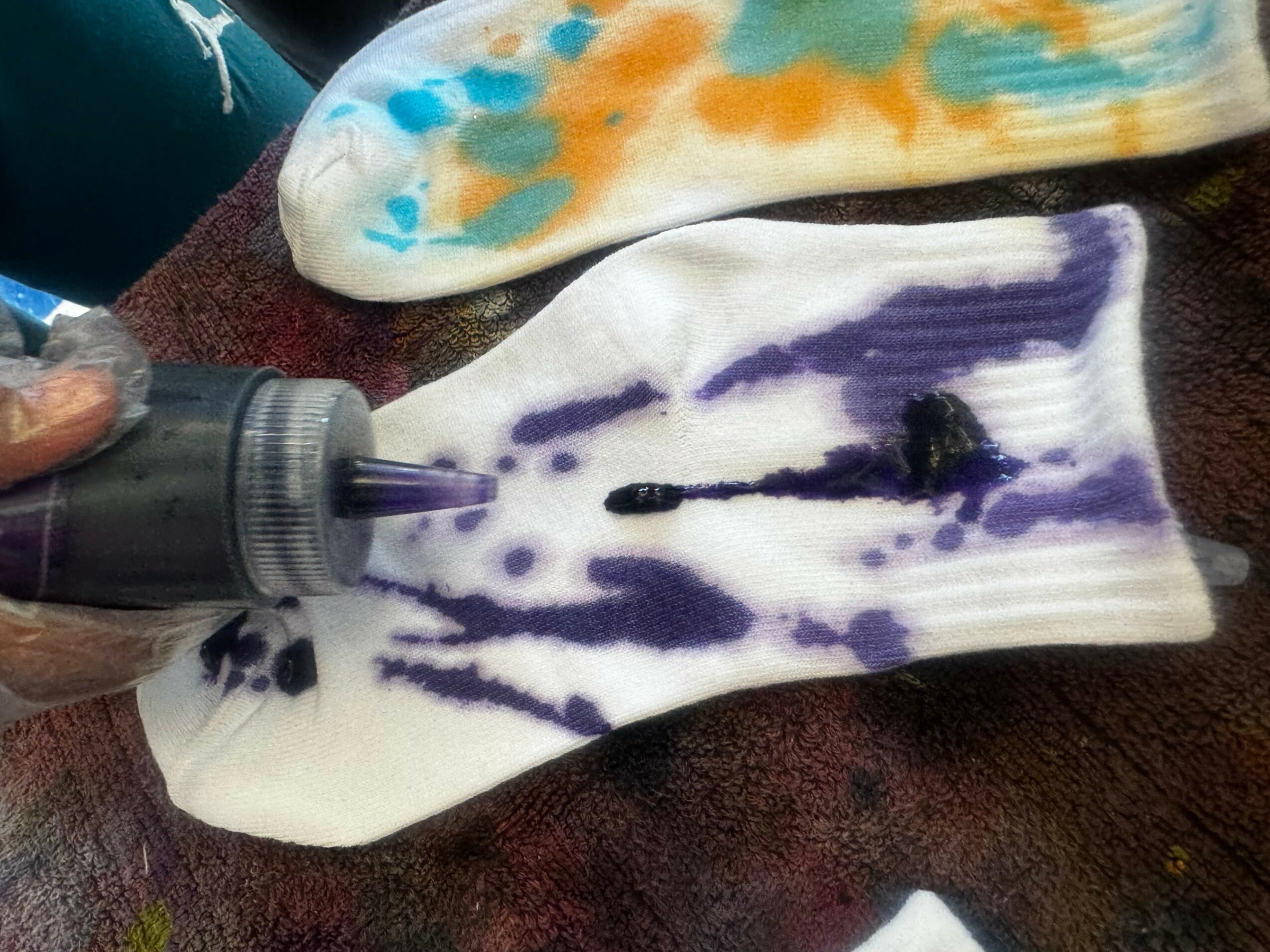 A sock is being dyed purple with a bottle of dye, with a colorful cloth nearby, on a dark surface.