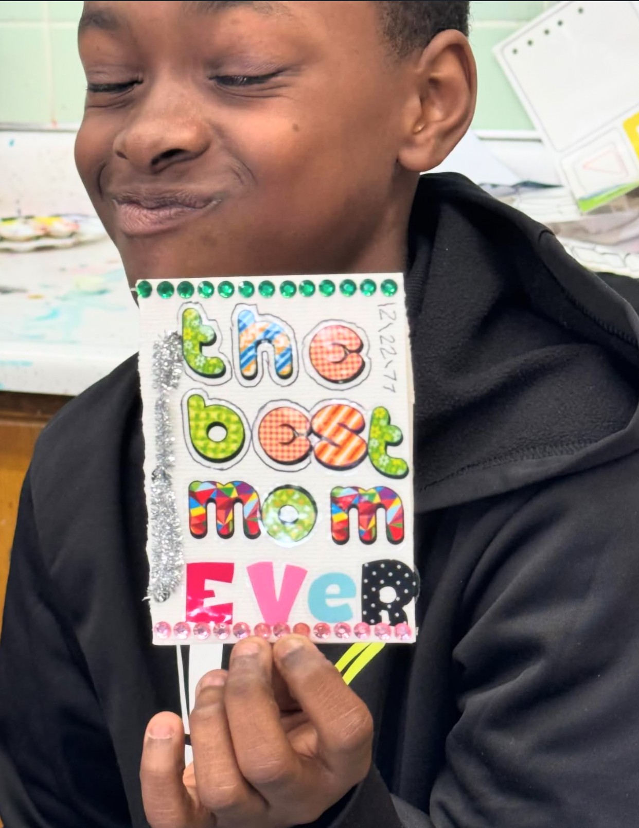 Child holding a colorful sign that says 'the best mom ever' with a classroom background.