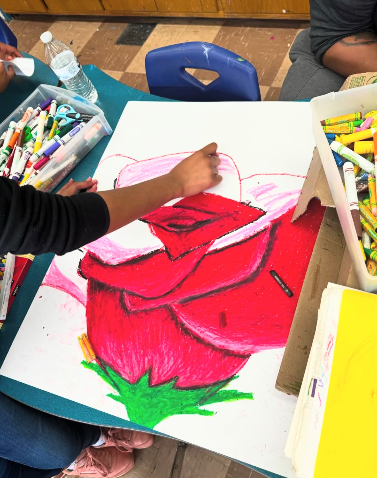 Child's hand drawing a large red rose on paper with markers, surrounded by art supplies, in a classroom setting.