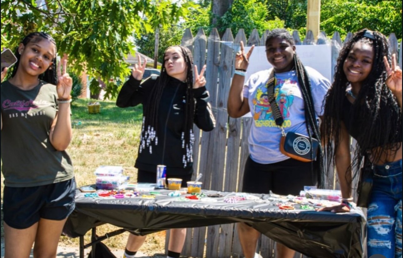 Four young women stand outdoors behind a table with various items, smiling and waving at the camera.