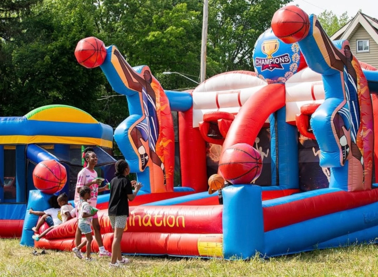 Children playing near a basketball-themed inflatable bounce house outdoors with trees and a house in the background.