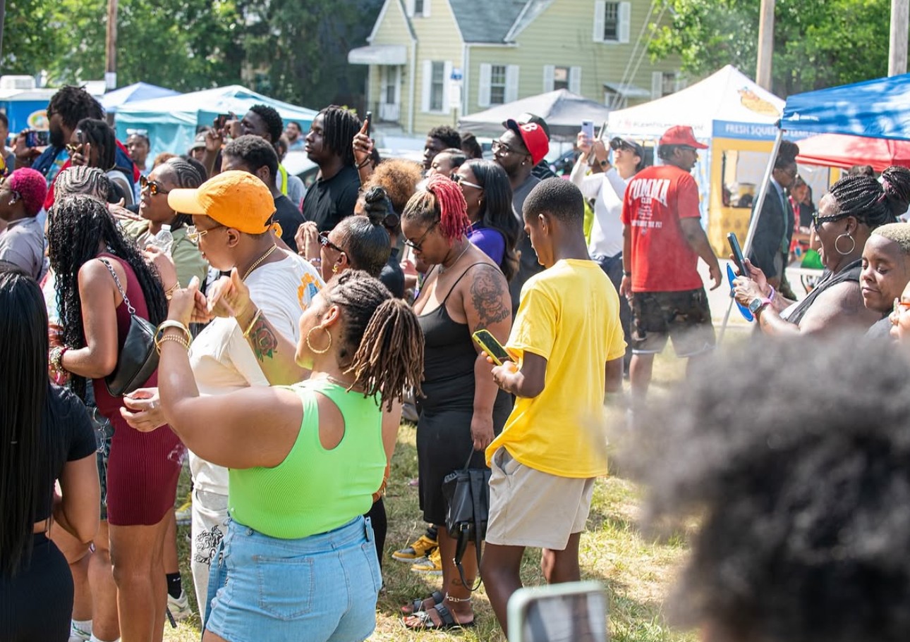 Crowd of people outdoors at a festival with tents and houses in background, some holding food or drinks.