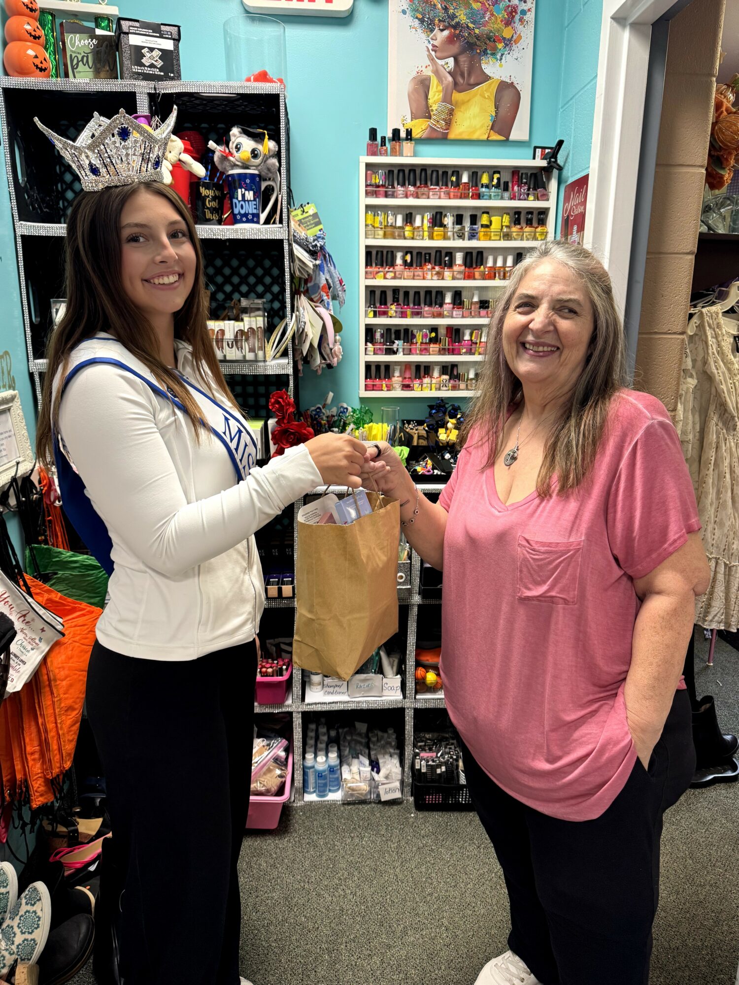 Two women shake hands inside a store, one wearing a crown and sash, the other in a pink top, smiling.