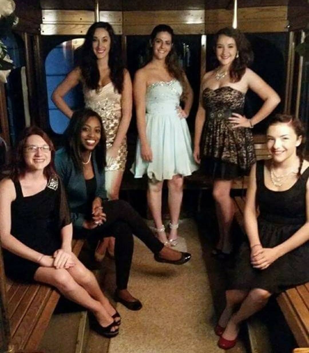 Group of seven women in dresses posing indoors, some sitting and some standing, smiling at the camera.