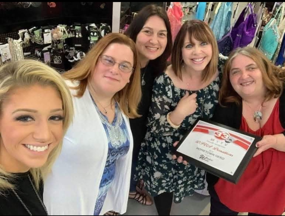 Group of five women smiling, holding a certificate, in a store with clothing in the background.
