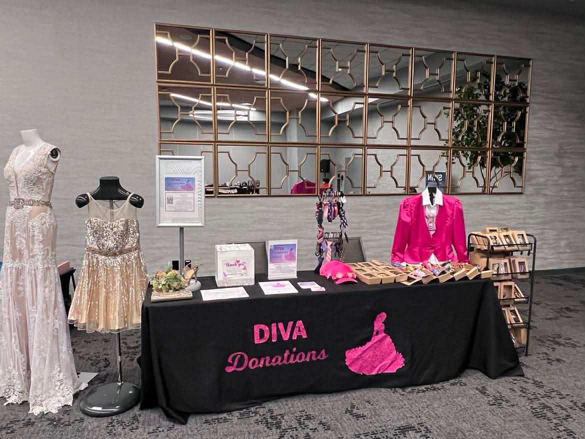 Table with pink dress and accessories, mannequins with dresses, decorative mirror, and donation sign, in a room with gray walls.