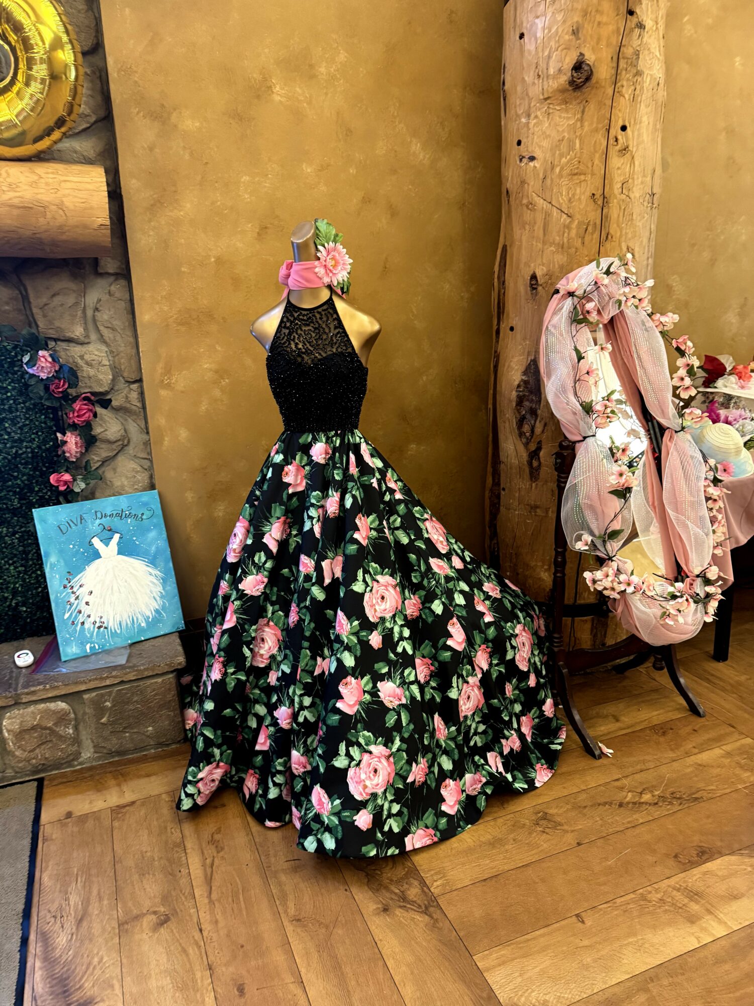 Decorative dress form with a floral dress and headpiece, surrounded by flowers and accessories, on a wooden floor.