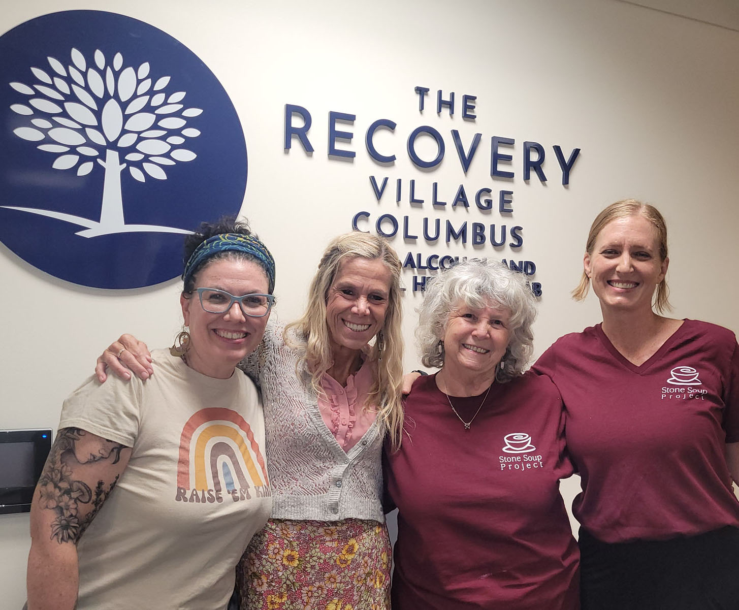 Four women standing together in front of a sign for The Recovery Village Columbus, smiling, with one woman wearing a rainbow shirt.