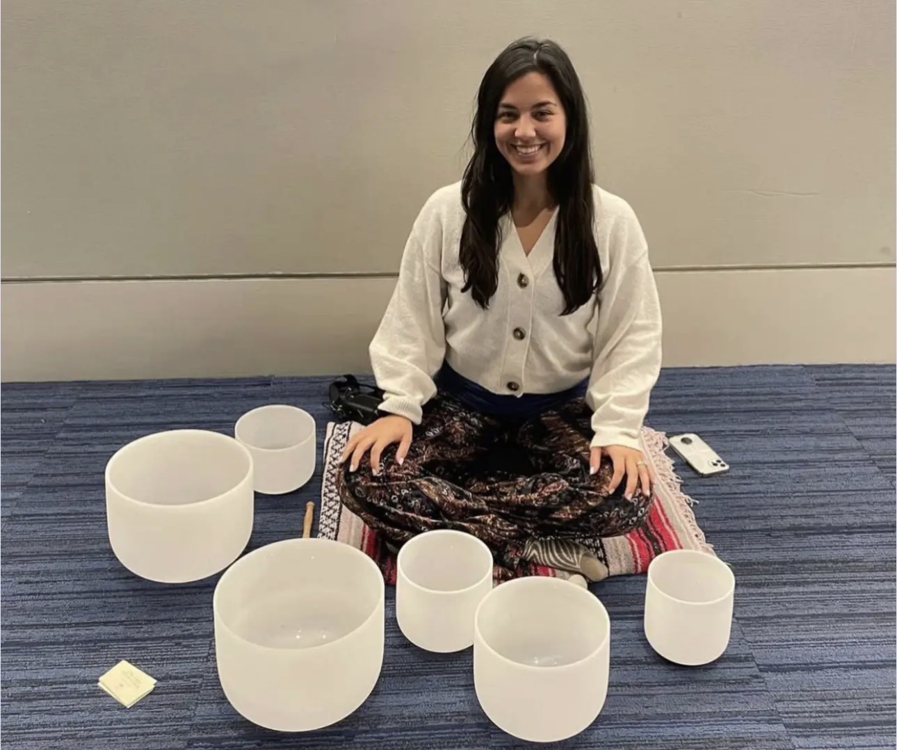 Young woman sitting on floor with singing bowls arranged in front of her, smiling, against a plain wall and blue carpet.
