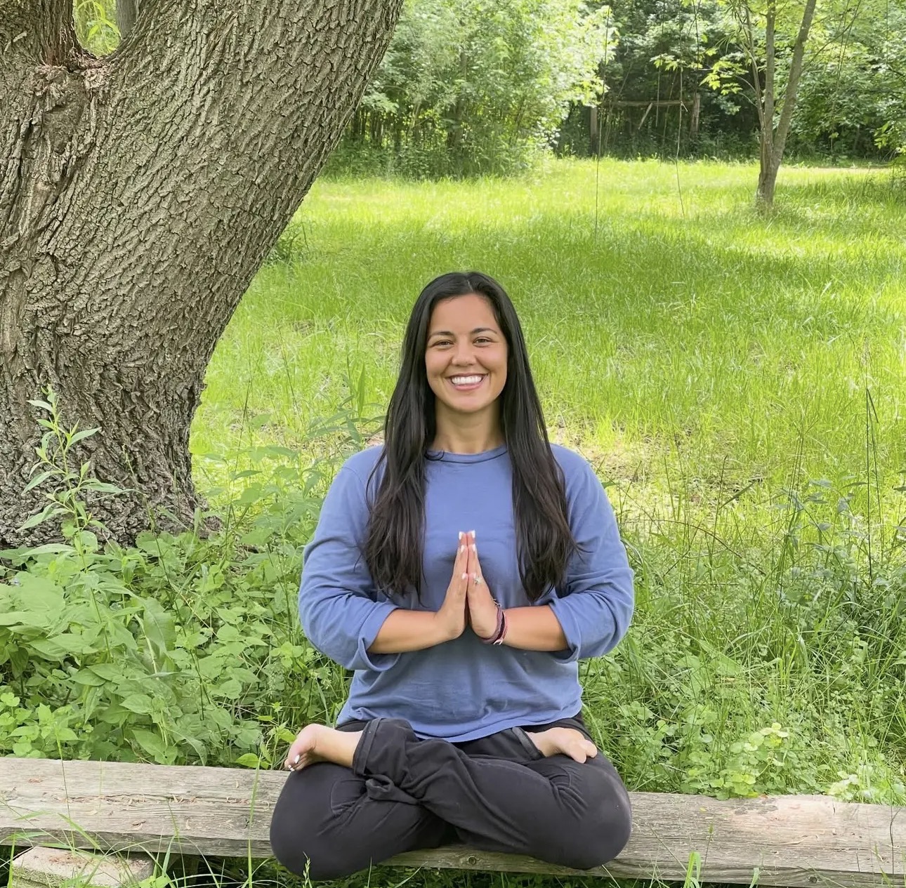 Person sitting cross-legged on a wooden bench outdoors, smiling with hands in prayer position, surrounded by green grass and trees.