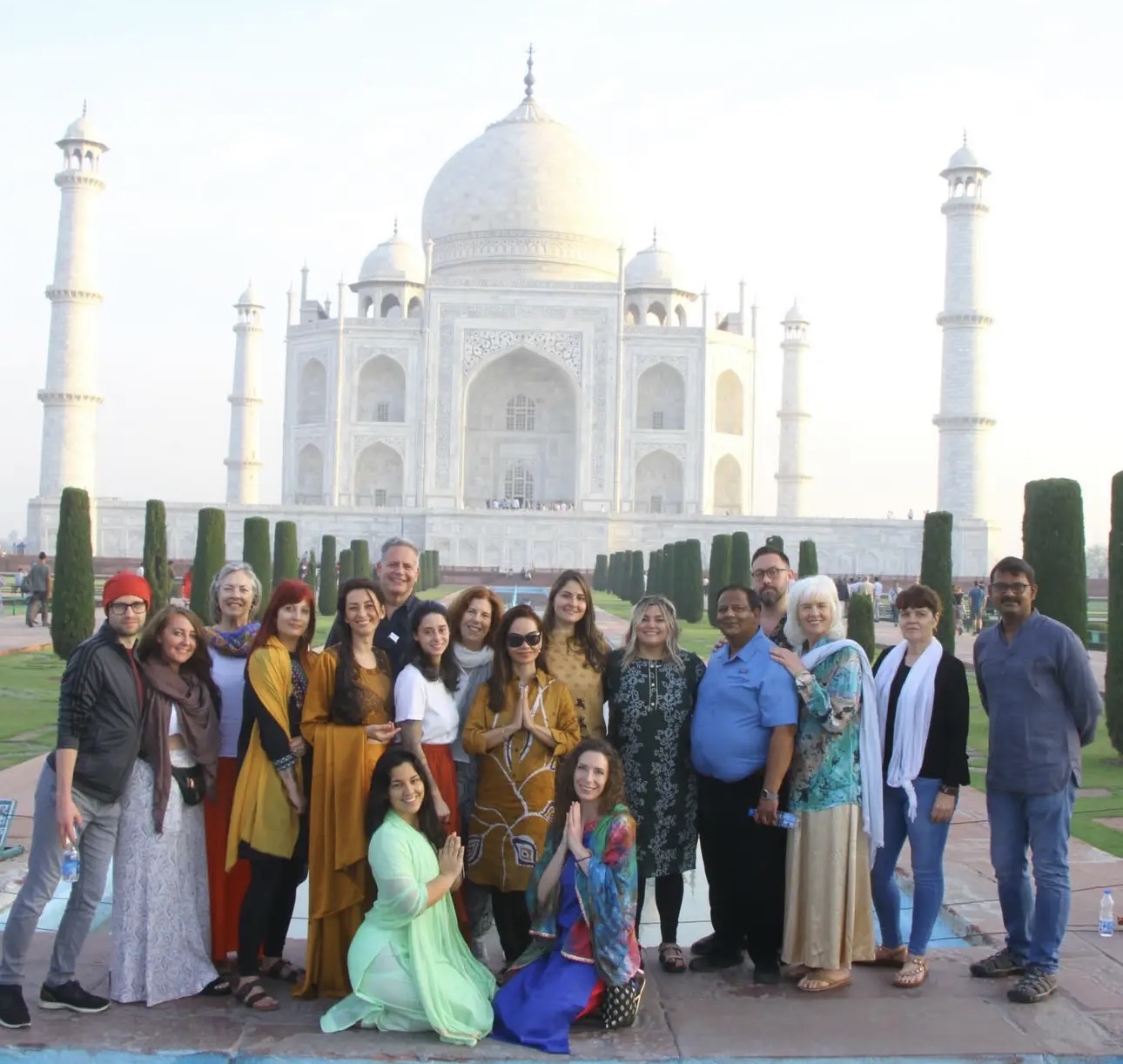 Group of people posing in front of the Taj Mahal, some smiling, with clear sky and gardens visible.