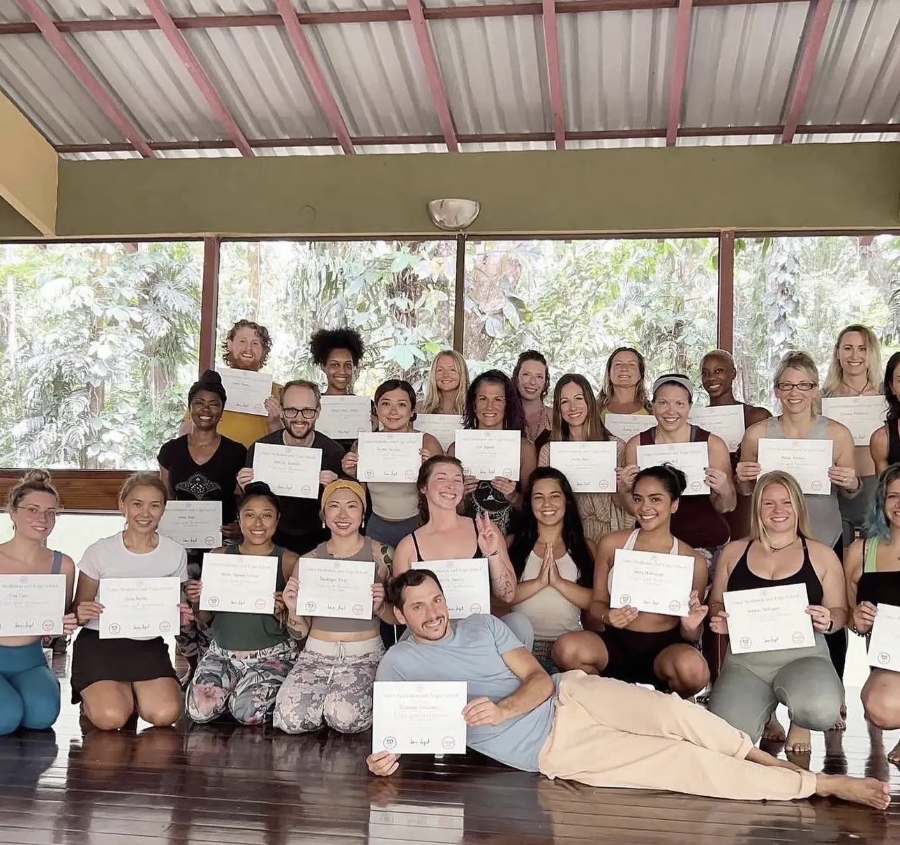 Group of people holding certificates indoors with large windows and trees outside, some sitting and some standing, one person lying in front.