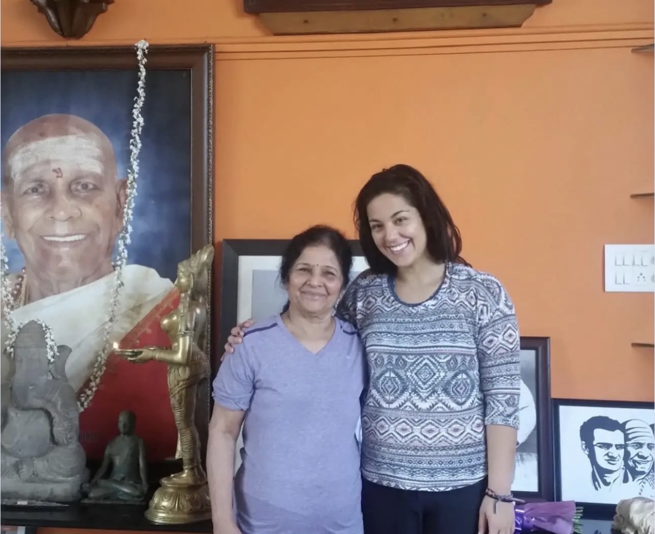 Two women standing indoors, smiling, with framed pictures and a statue behind them.