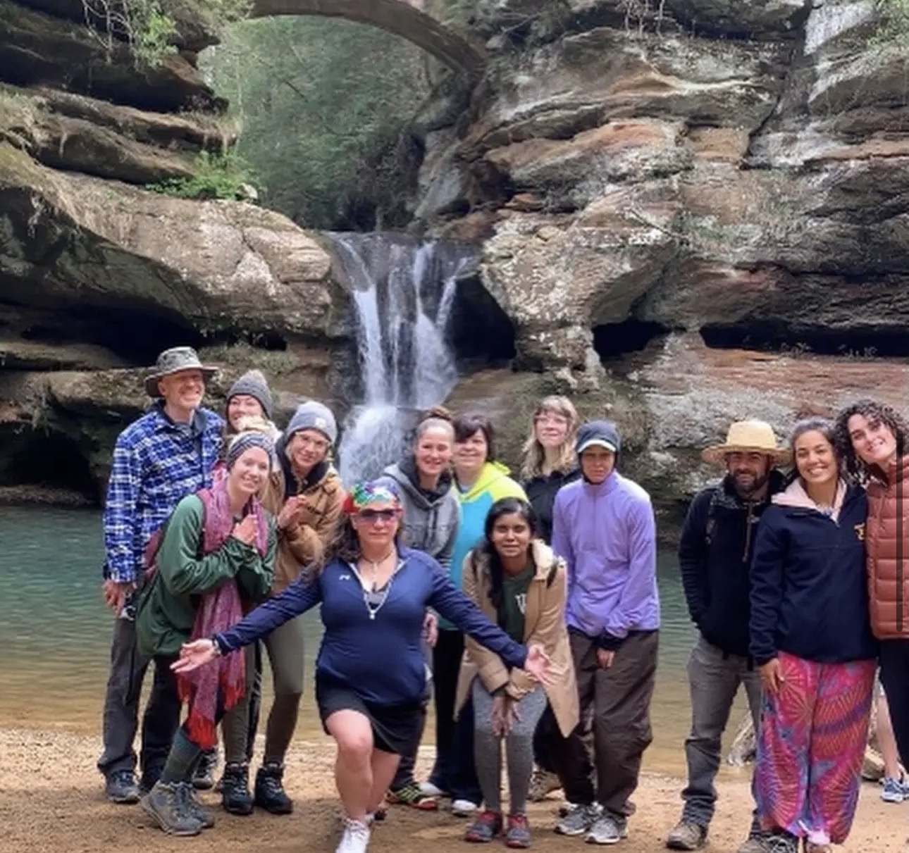 Group of people posing outdoors near a waterfall with rocks and trees in the background.