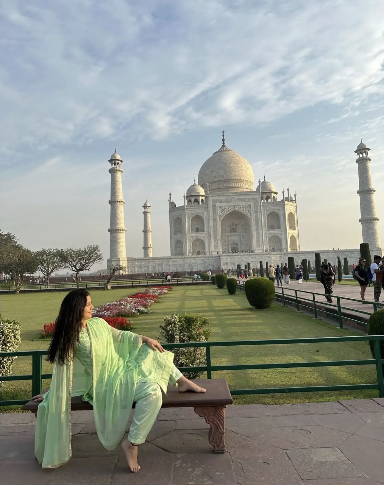 Woman in green dress sitting on a bench in front of the Taj Mahal, with a garden and visitors nearby.