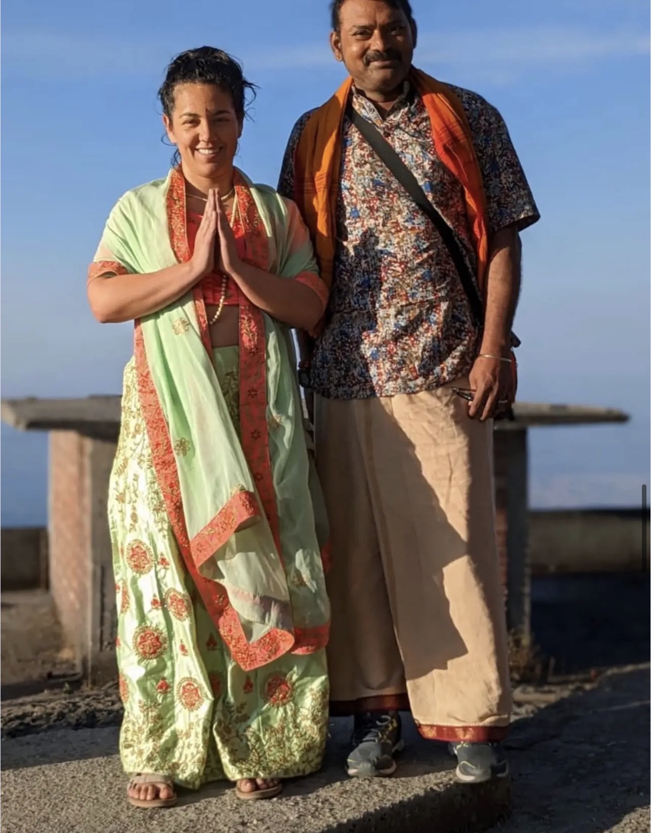 Two people standing outdoors near water, smiling, with hands in a greeting gesture, wearing traditional clothing, under a blue sky.