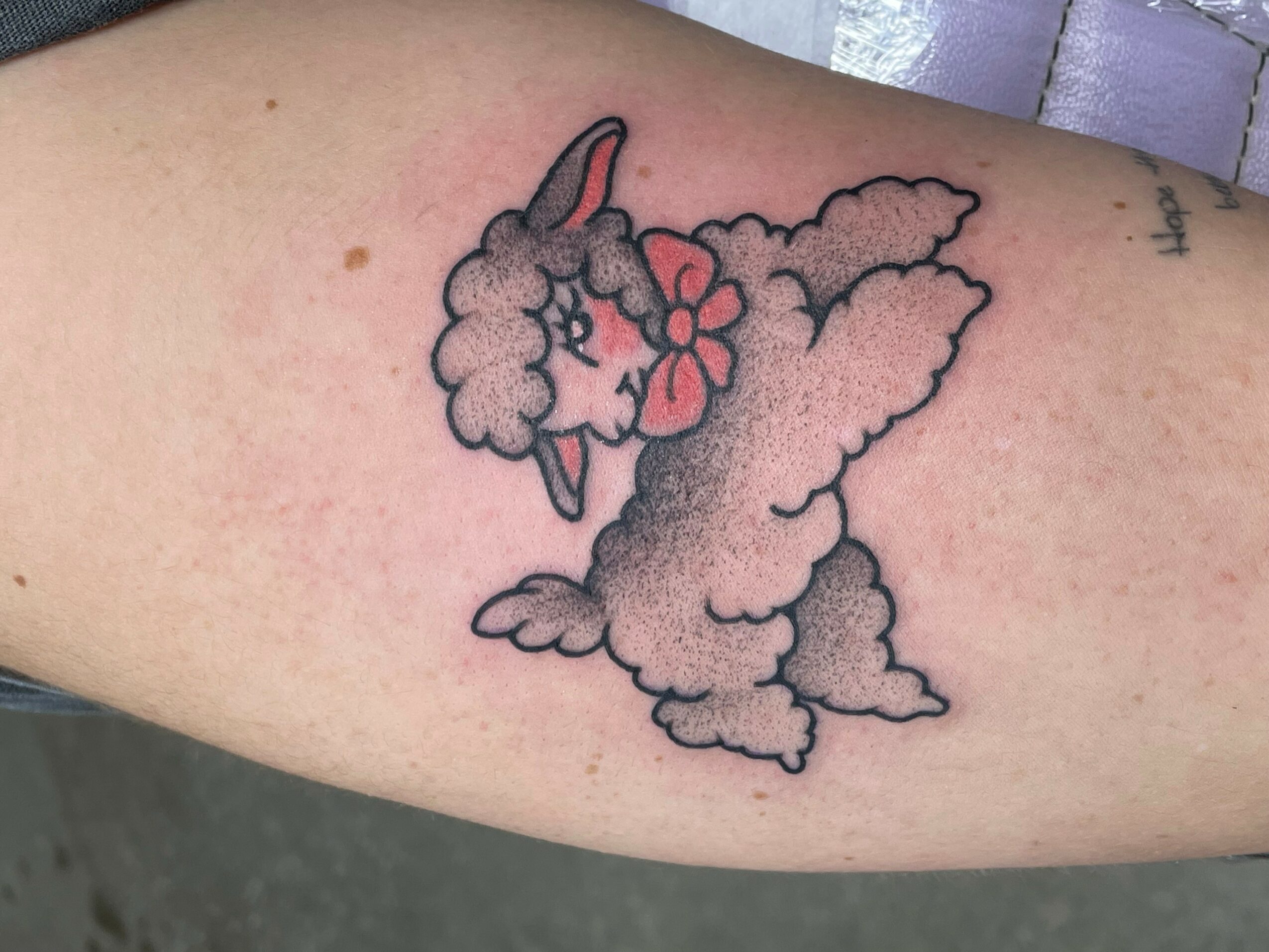 Tattoo of a cartoon unicorn with a pink bow, closed eyes, and a fluffy body on skin.