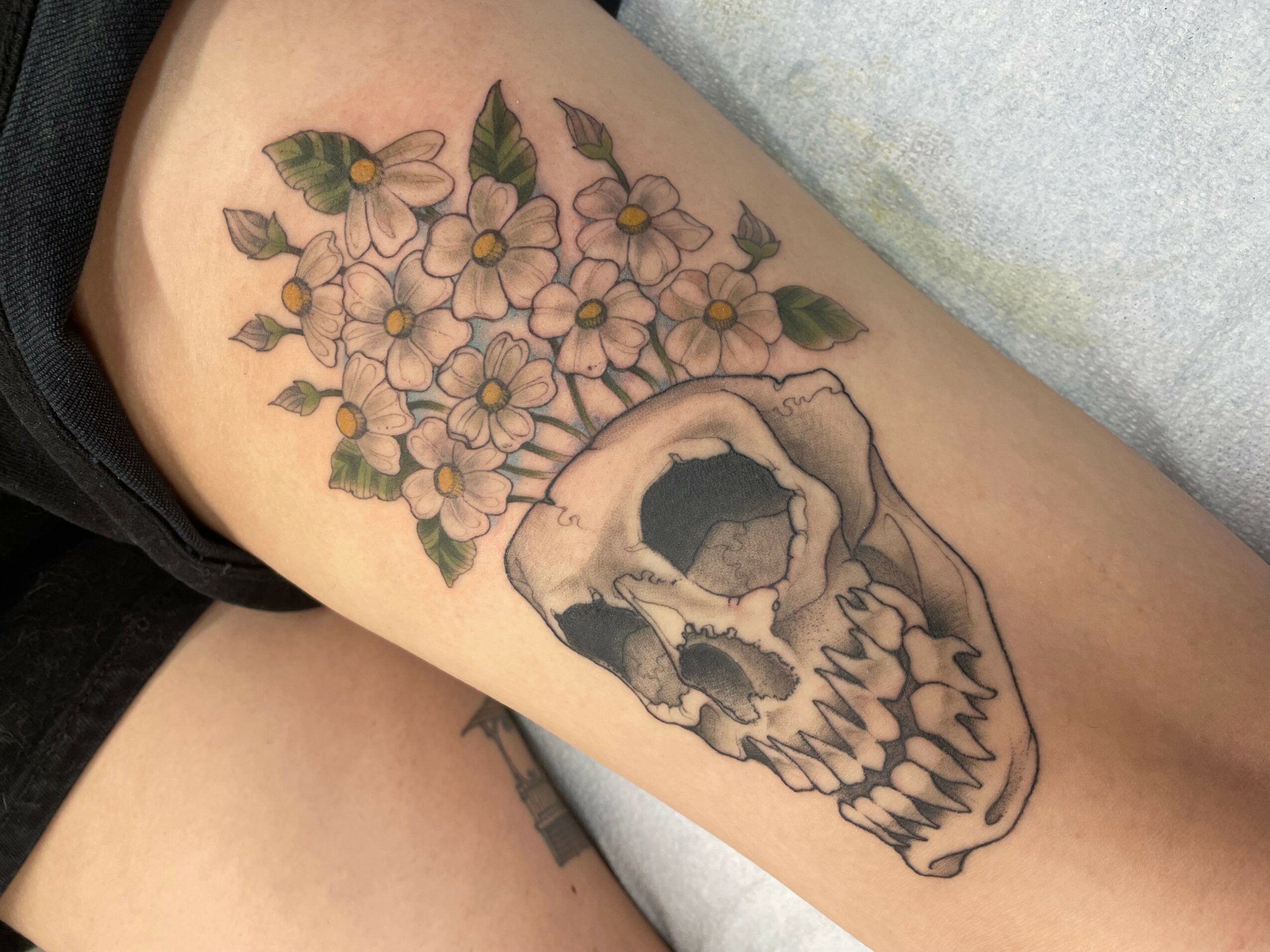 Tattoo of a skull surrounded by pink flowers and green leaves on a person's arm.