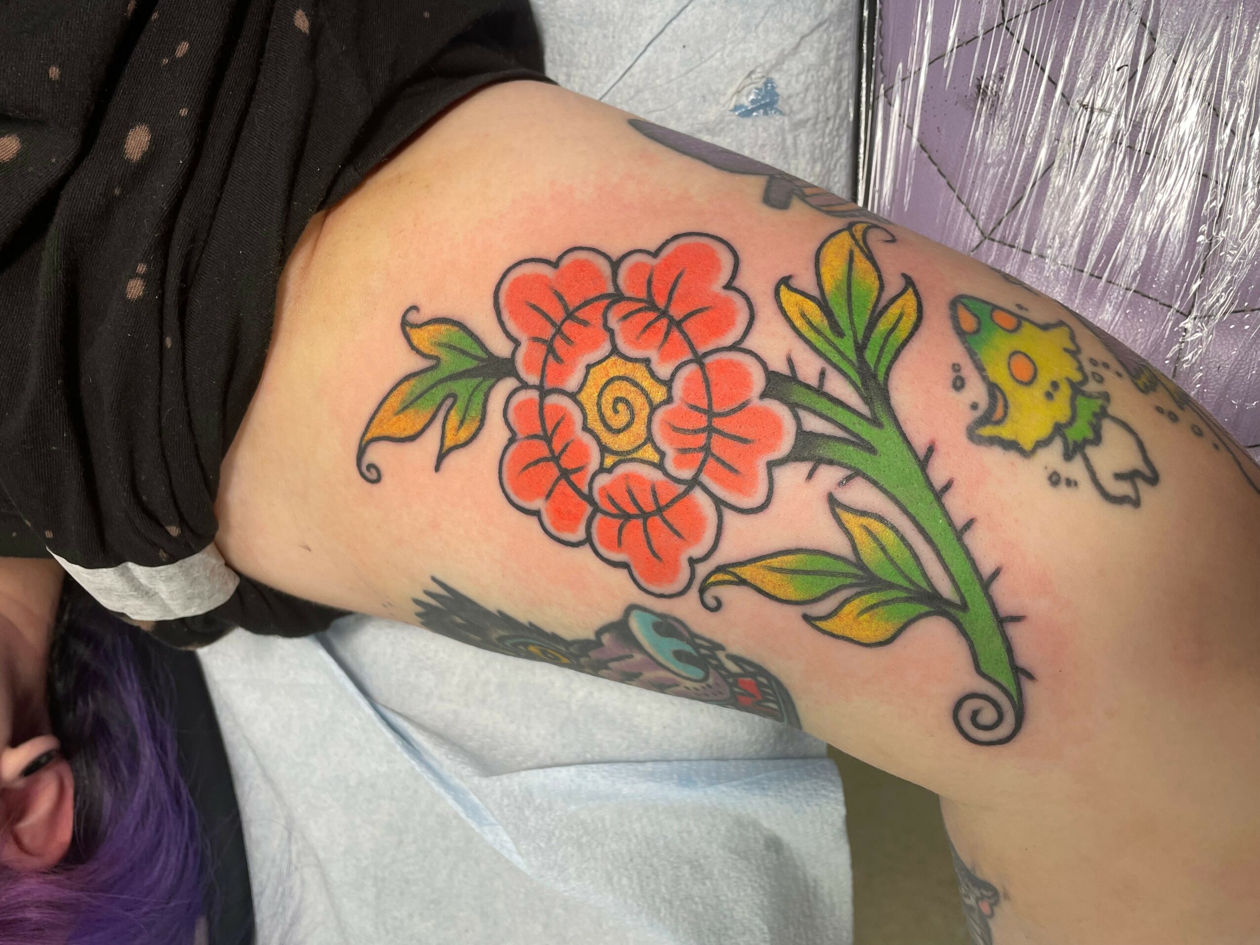 Tattoo of a large red flower with green leaves on a person's thigh, with additional smaller tattoos nearby.