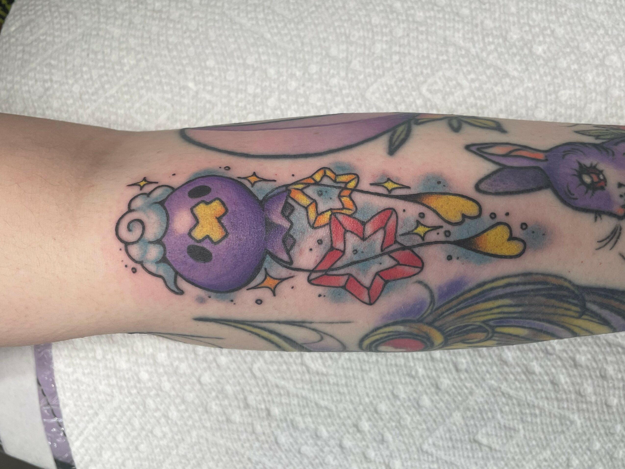 Colorful tattoo of a purple bomb with a yellow cross, surrounded by stars, clouds, and abstract shapes on a person's arm.
