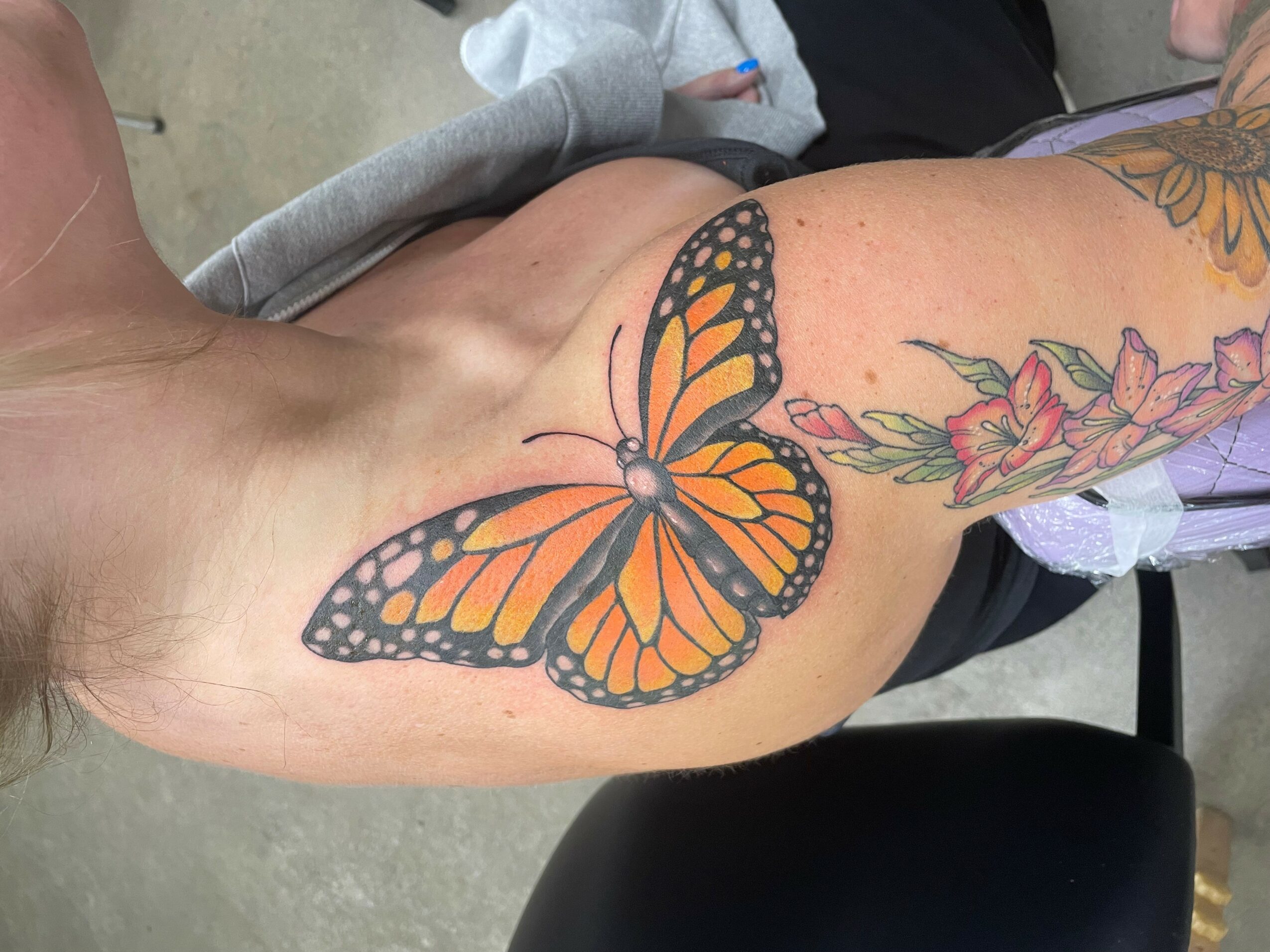 Close-up of a butterfly tattoo on a person's shoulder and upper arm, with colorful flowers nearby.
