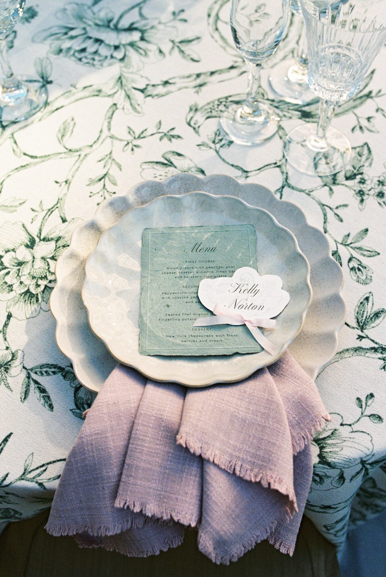 A scalloped-edged plate with a menu, a heart-shaped note, and a pink cloth on a floral tablecloth.