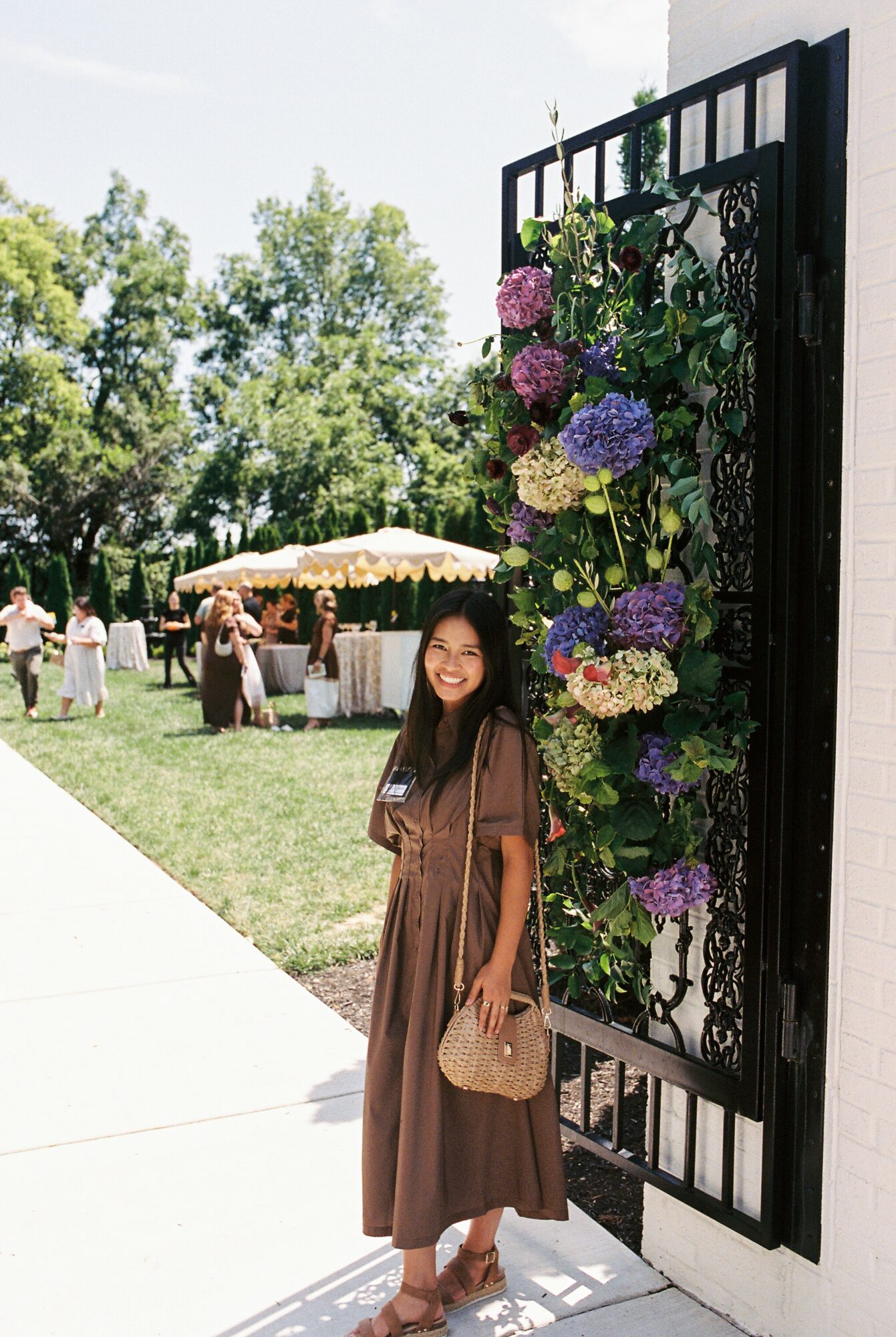 Woman in brown dress smiling next to a large floral display on a black gate outdoors, with tents and trees in background.