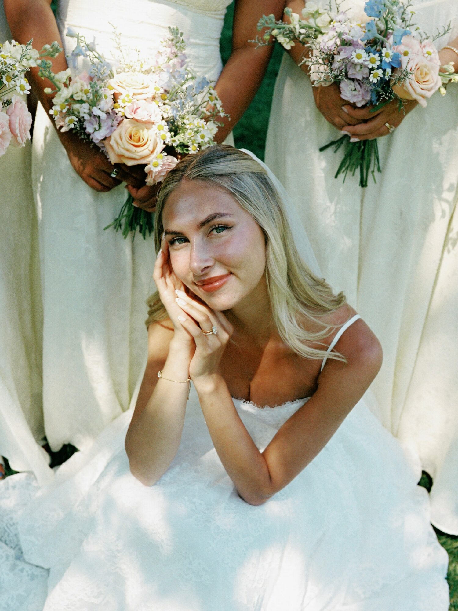 Young woman in a white dress sitting on the ground, smiling with hands near her face, surrounded by women holding bouquets.