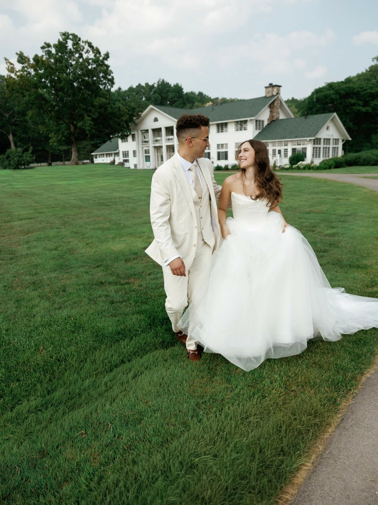 A bride and groom walk on grass near a large white house, holding hands and looking at each other.
