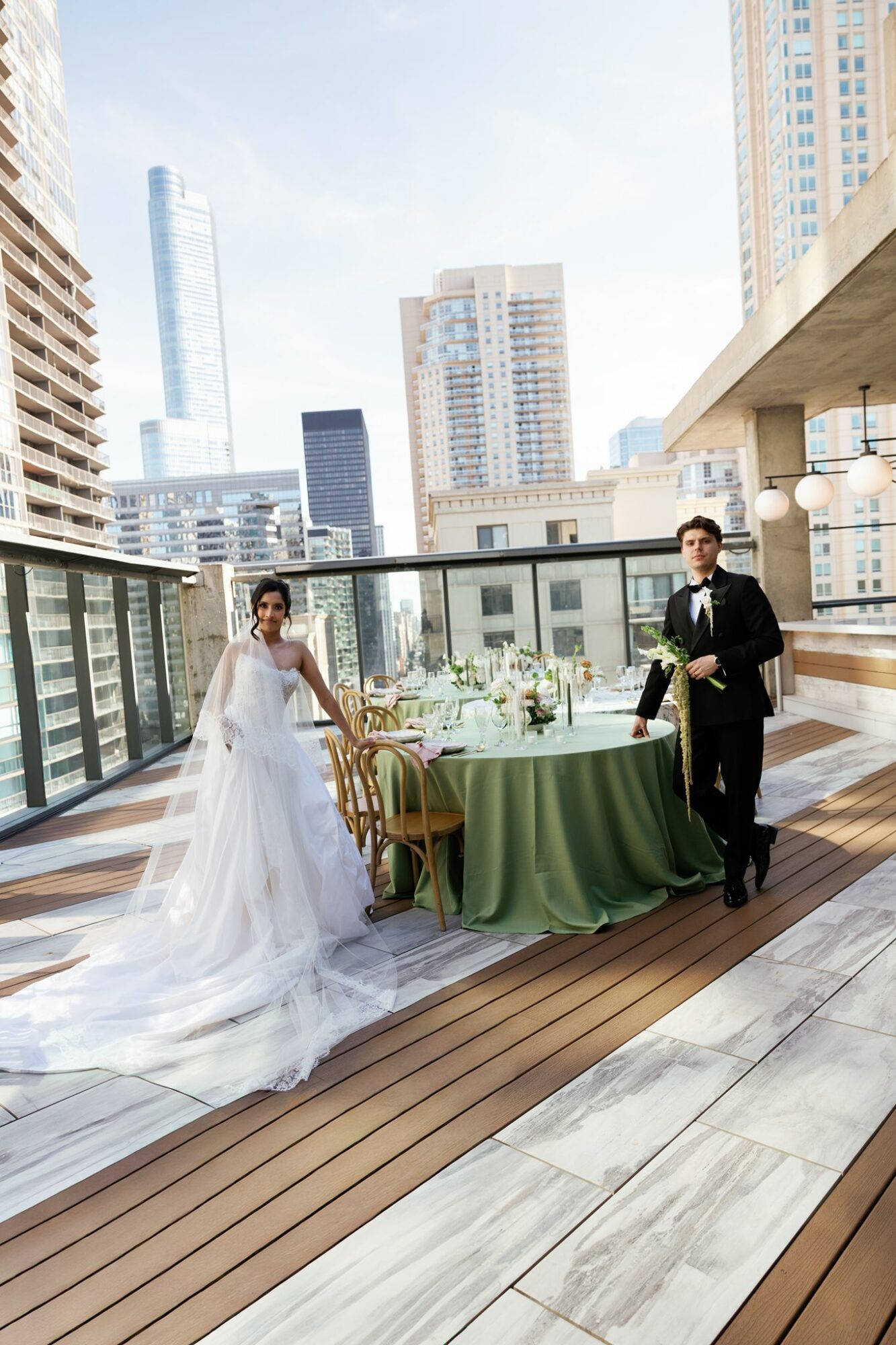Bride and groom stand beside a decorated table on a rooftop with city skyscrapers in the background, sunny sky.