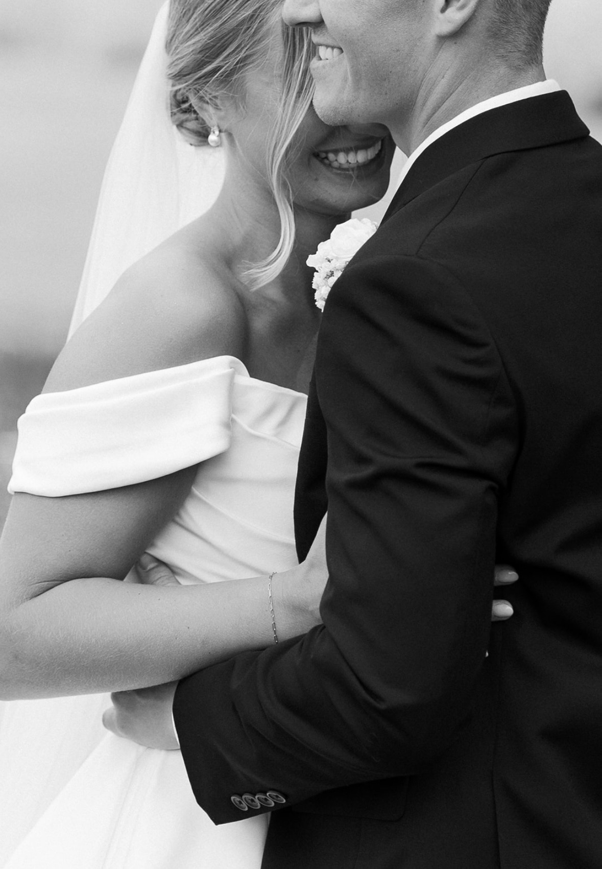 A bride and groom embrace closely, smiling, with the bride wearing an off-shoulder wedding dress and veil, and the groom in a suit.