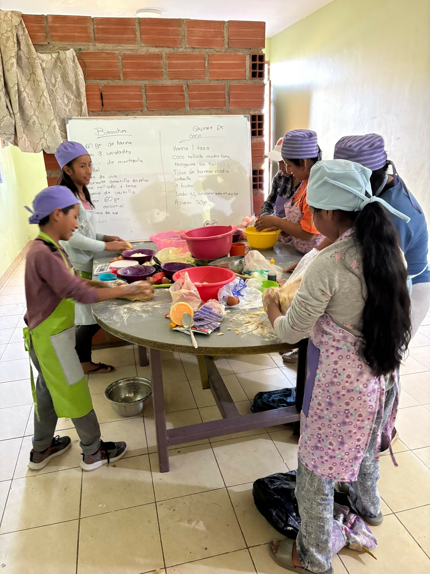 People gathered around a table with food and drinks, some wearing masks, in an indoor setting with brick wall and whiteboard.