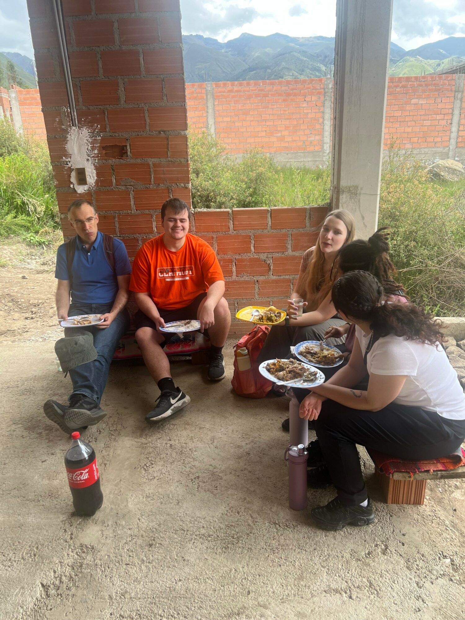 Three people sitting on a small bench outside, eating food, with a brick wall and mountains in the background.