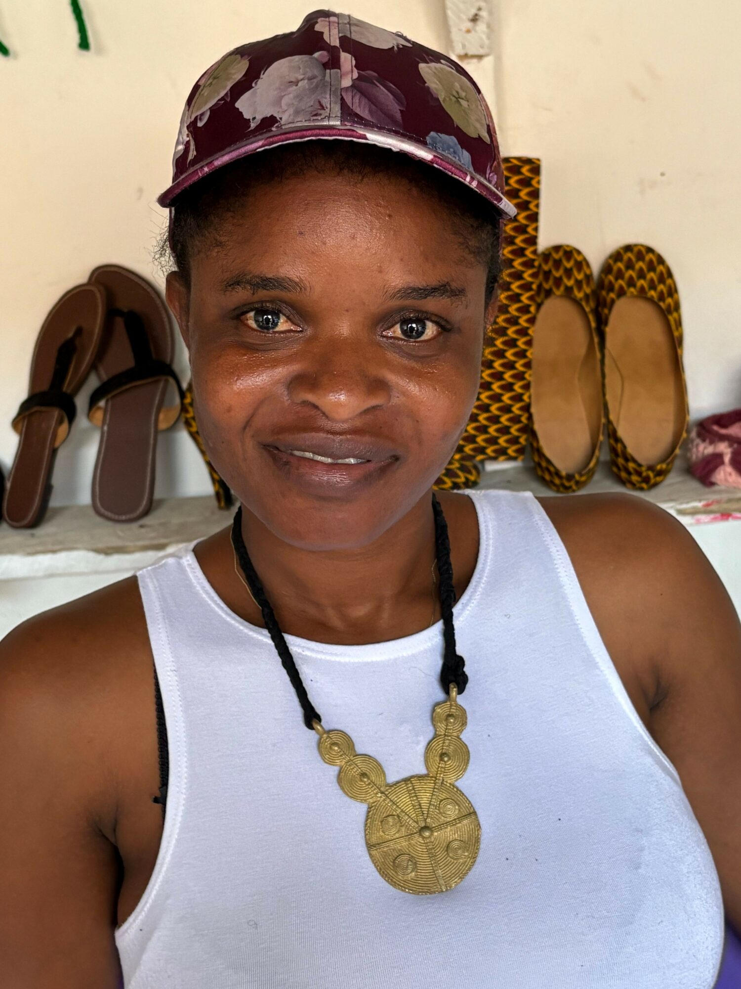 Woman wearing a white tank top, patterned hat, and a large necklace, standing outdoors with shoes and bags in the background.