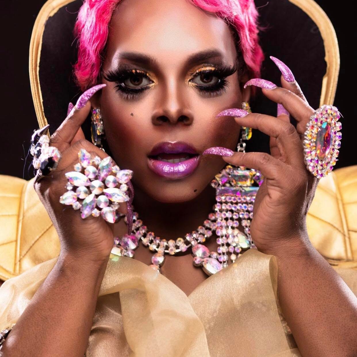 Woman with pink hair, wearing jewelry, holding her face with both hands, looking directly at the camera.