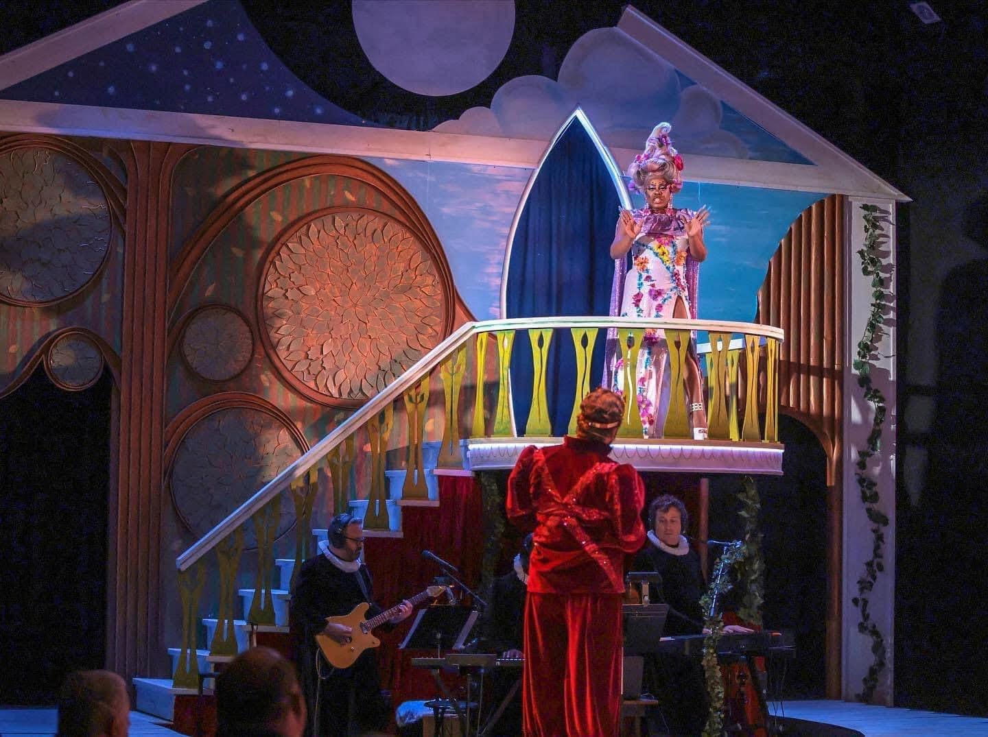 Stage with a performer in colorful costume on a balcony, musicians playing, and audience in front.