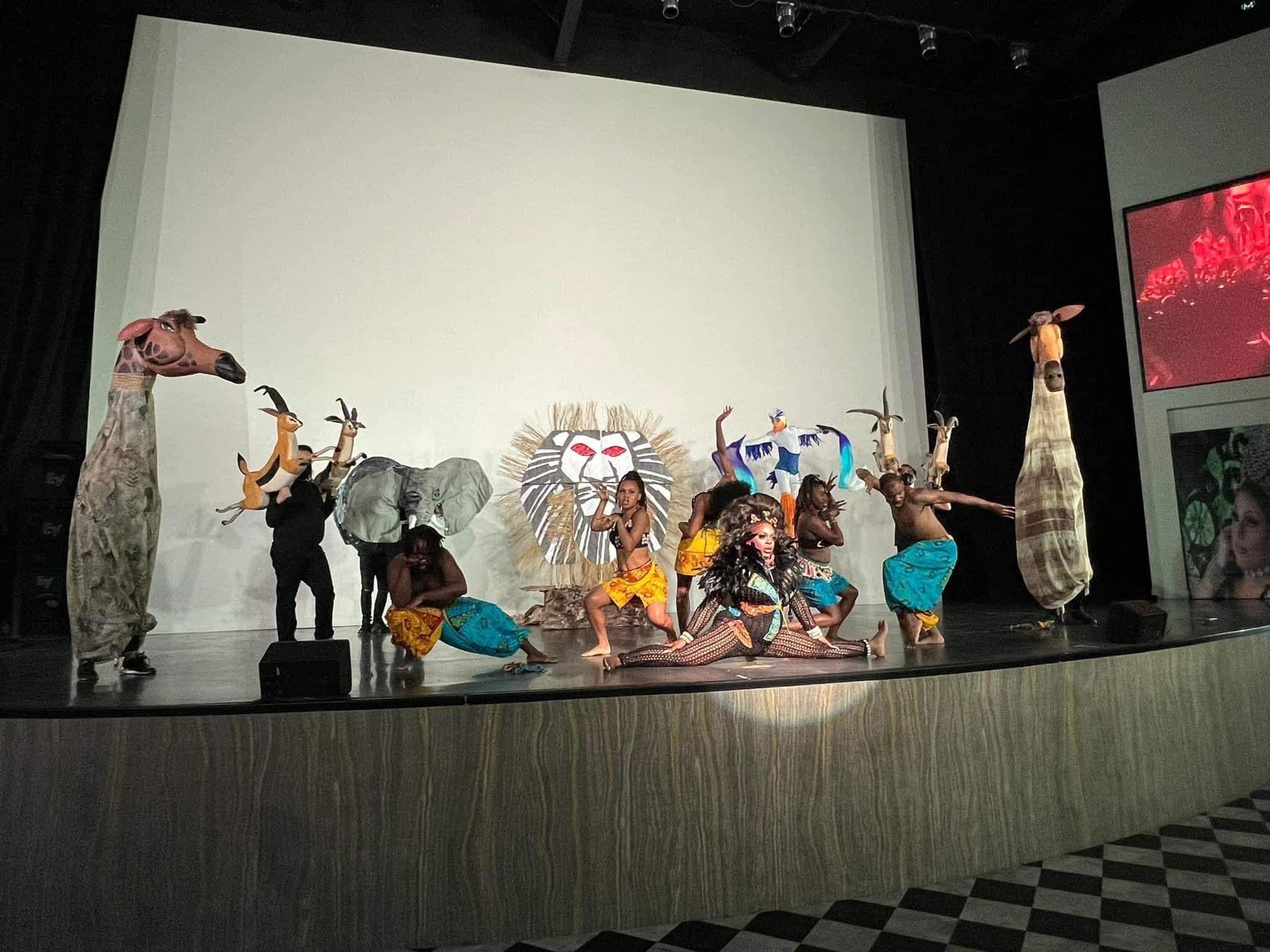 Children in costumes perform on stage with animal and tribal-themed props, against a plain white background.