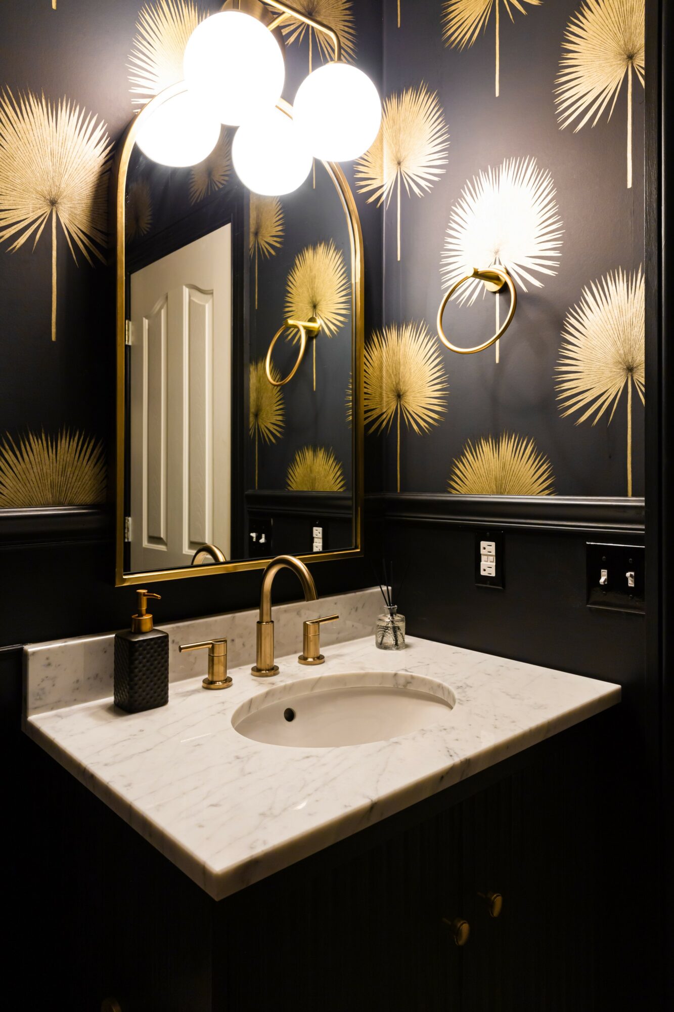 Bathroom vanity with a white marble countertop, gold fixtures, and a mirror, against a dark wallpaper with gold fan patterns.