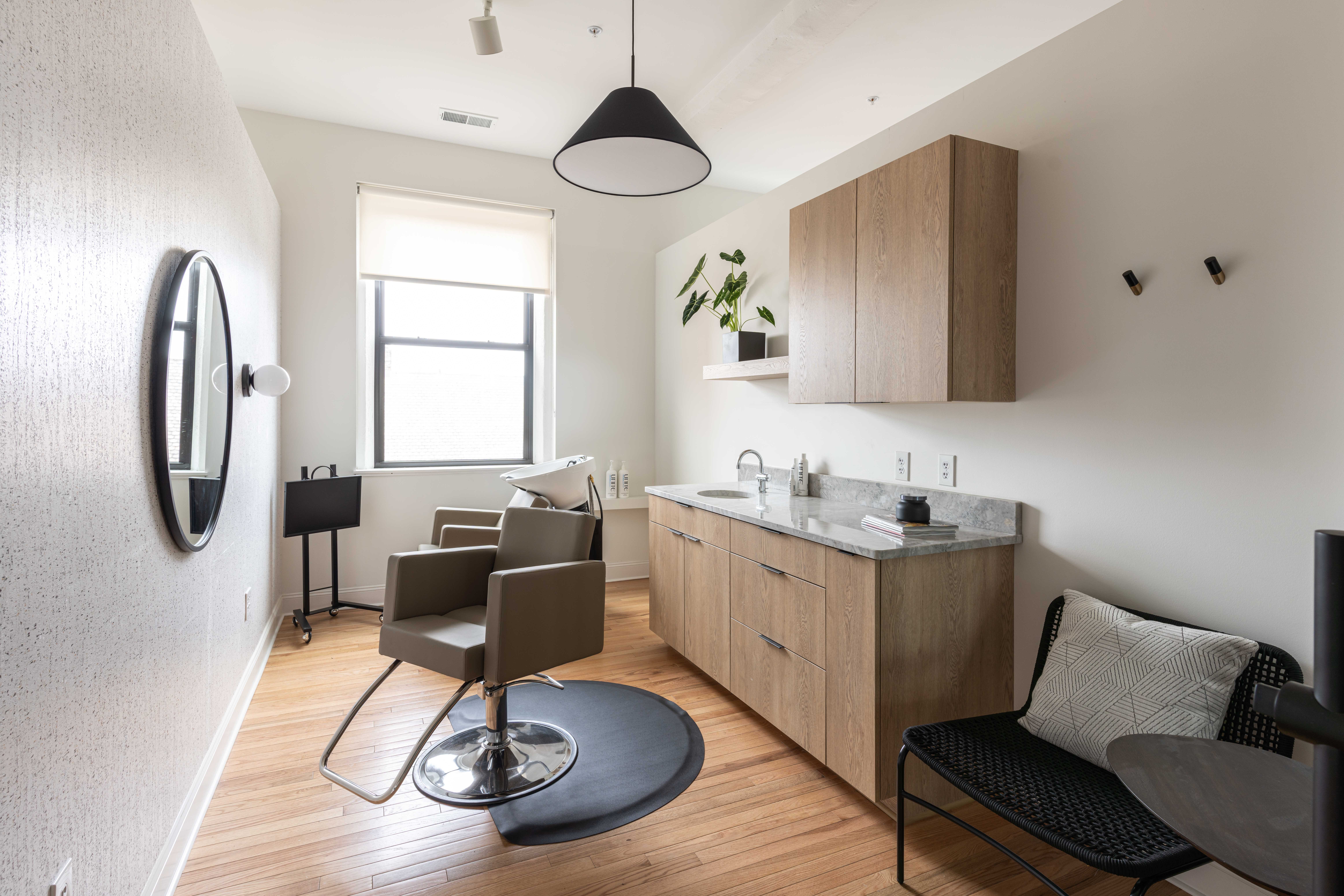 Small kitchen with a window, wooden cabinets, a sink, a chair, and a small table, with a black pendant light and wall hooks.
