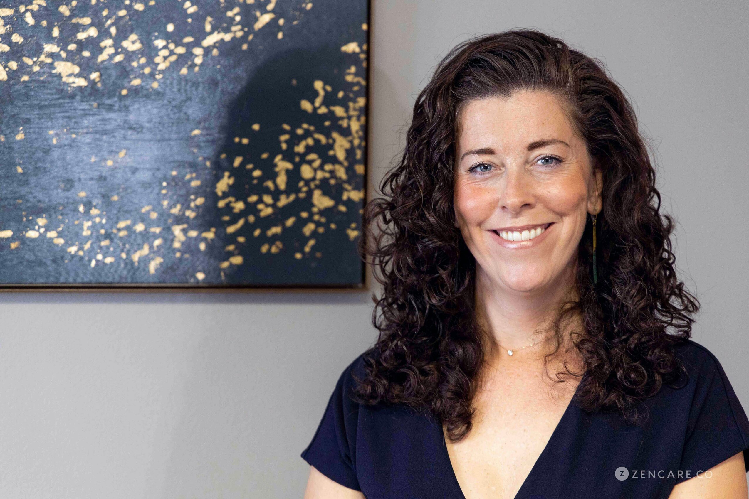 Woman with curly dark hair smiling, wearing a dark top, standing indoors with abstract art on the wall behind her.