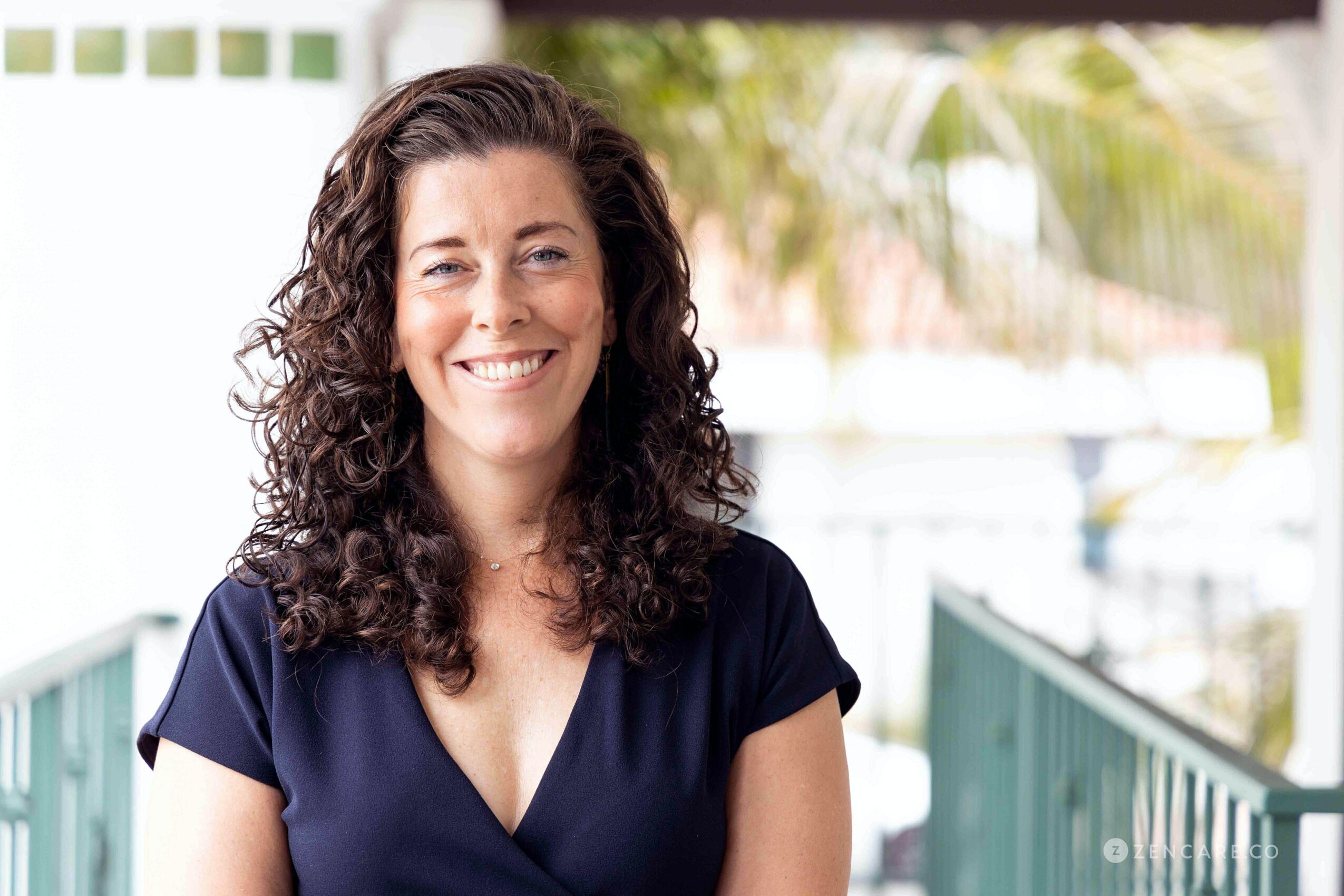 Smiling woman with curly hair wearing a navy top outdoors, with a blurred background of trees and a railing.