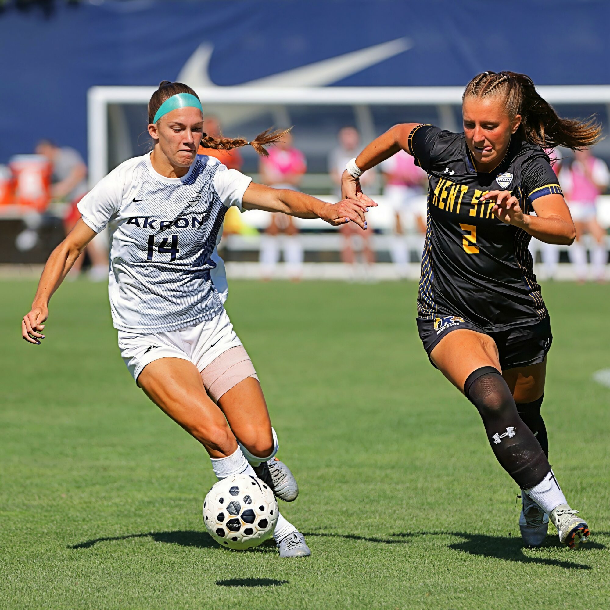 Two female soccer players compete for the ball on a green field, one in white and one in black uniform.