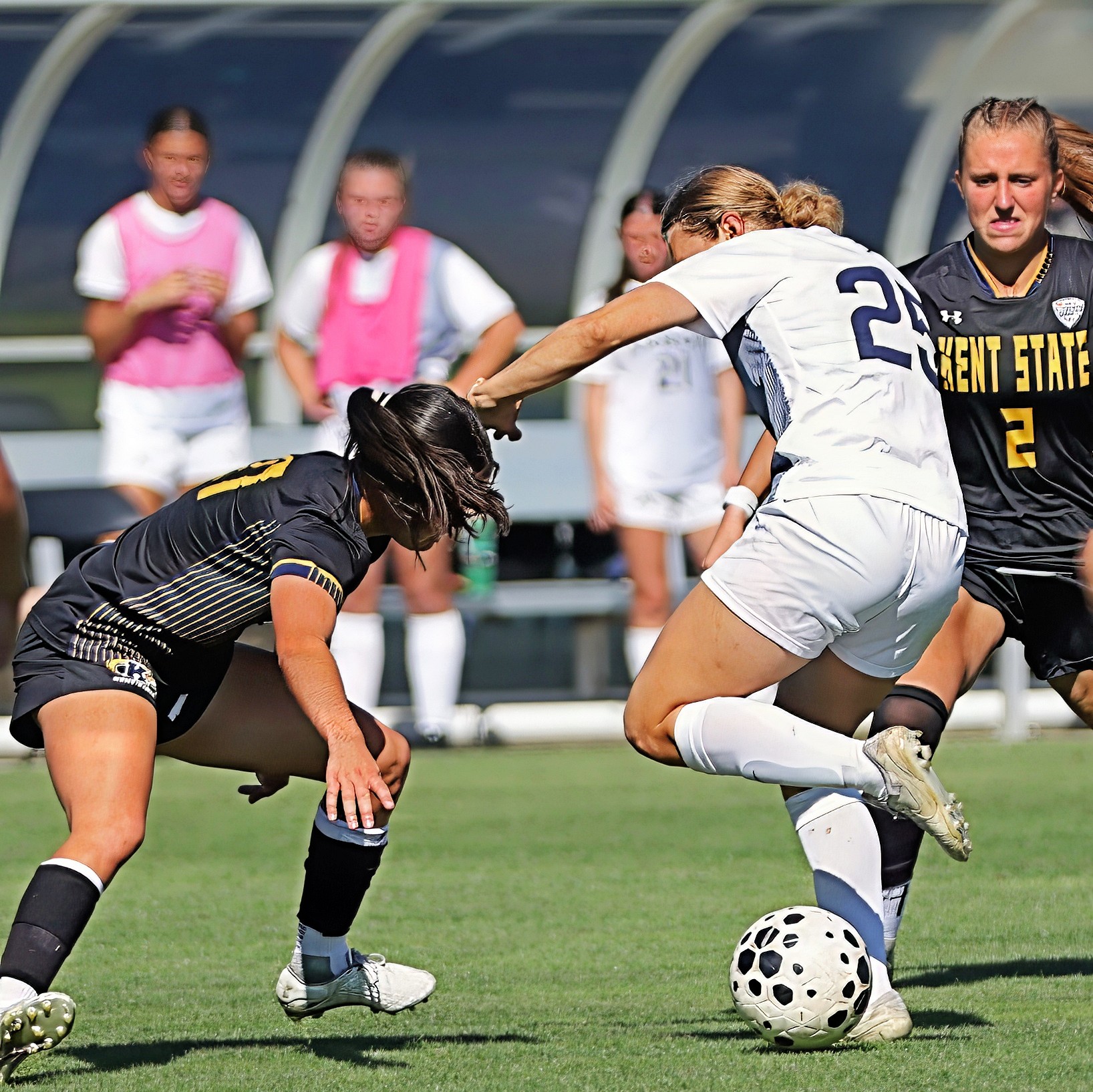 Two female soccer players compete for the ball on the field, with three players and a dugout in the background.