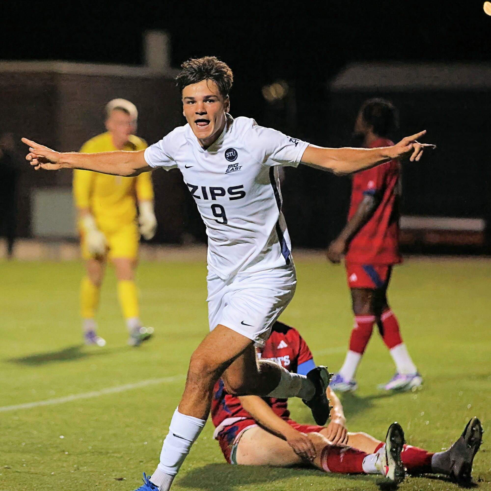 Soccer player celebrating with arms outstretched on field, other players in background, nighttime setting