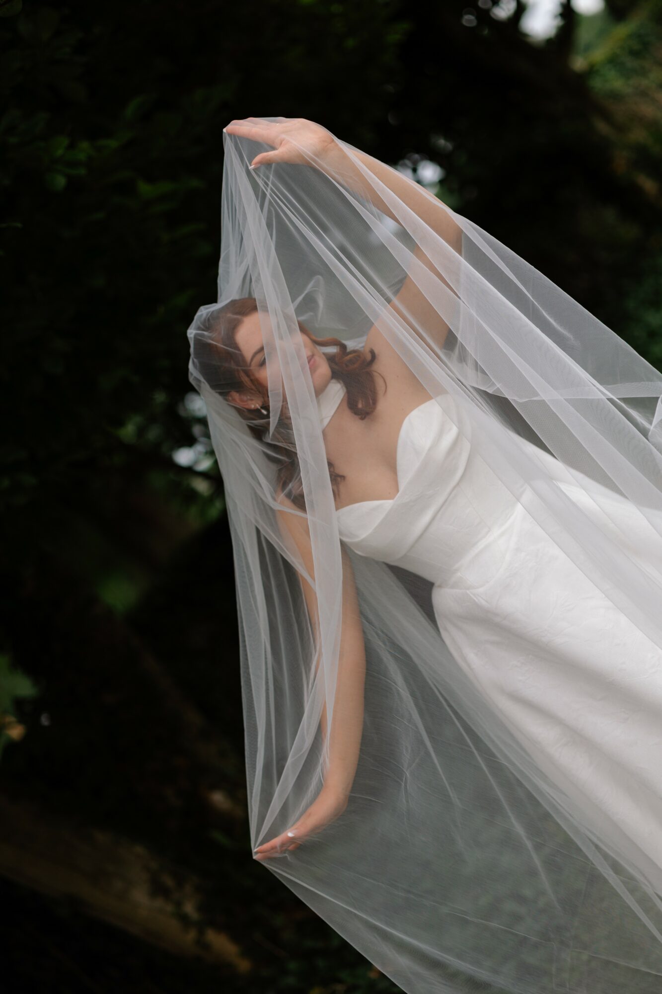 Bride with veil outdoors, holding veil above her head, wearing a strapless white dress, dark hair, trees in background.