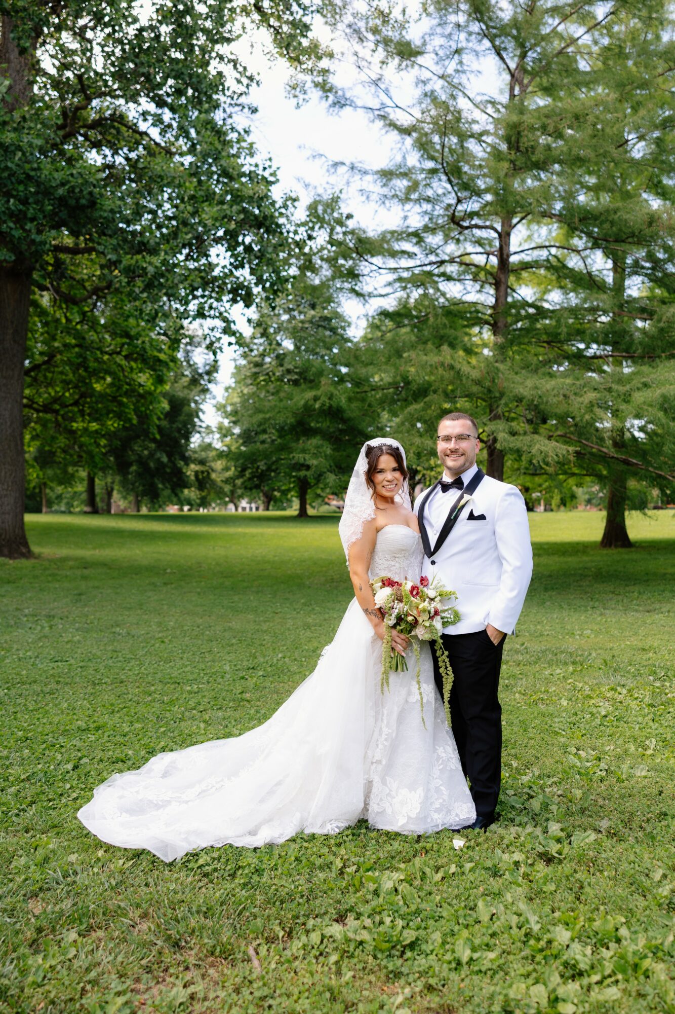 Bride and groom standing on grass in a park with trees, smiling, bride holding a bouquet, bride in a white gown, groom in a tuxedo.
